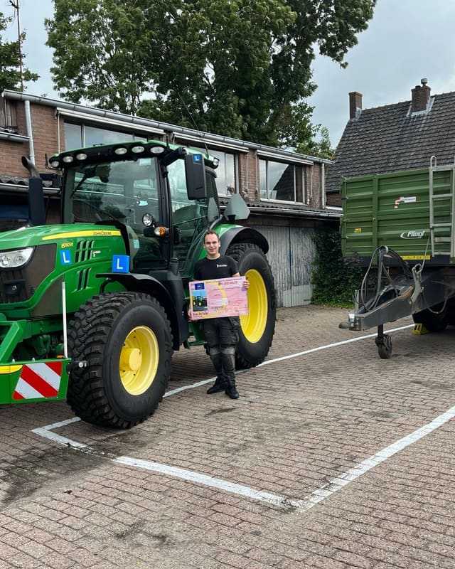 Geslaagde cursist met rijbewijsbord naast een groene lestrekker en een lege aanhanger