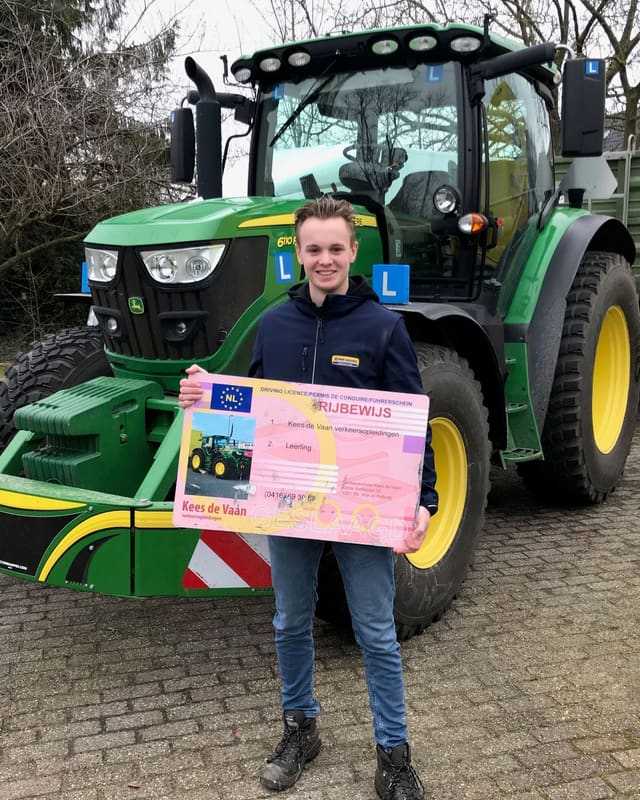 Geslaagd voor het trekkerrijbewijs: lachende cursist met rijbewijsbord voor de bumper van een groene lestrekker