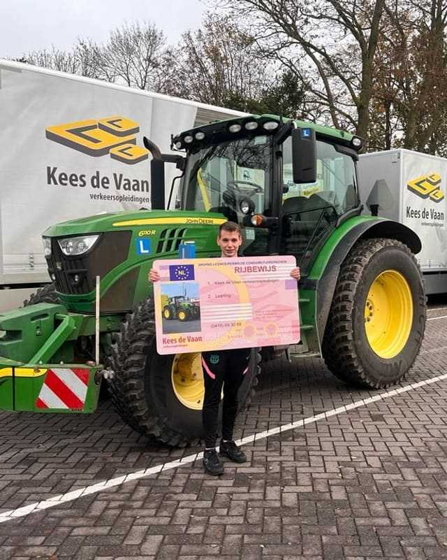 Geslaagd voor het trekkerrijbewijs: cursist met rijbewijsbord voor de lestrekker, met lesvrachtwagens op de achtergrond